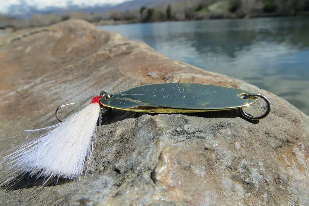 gold kastmaters fishing lure sitting on a rock by a river