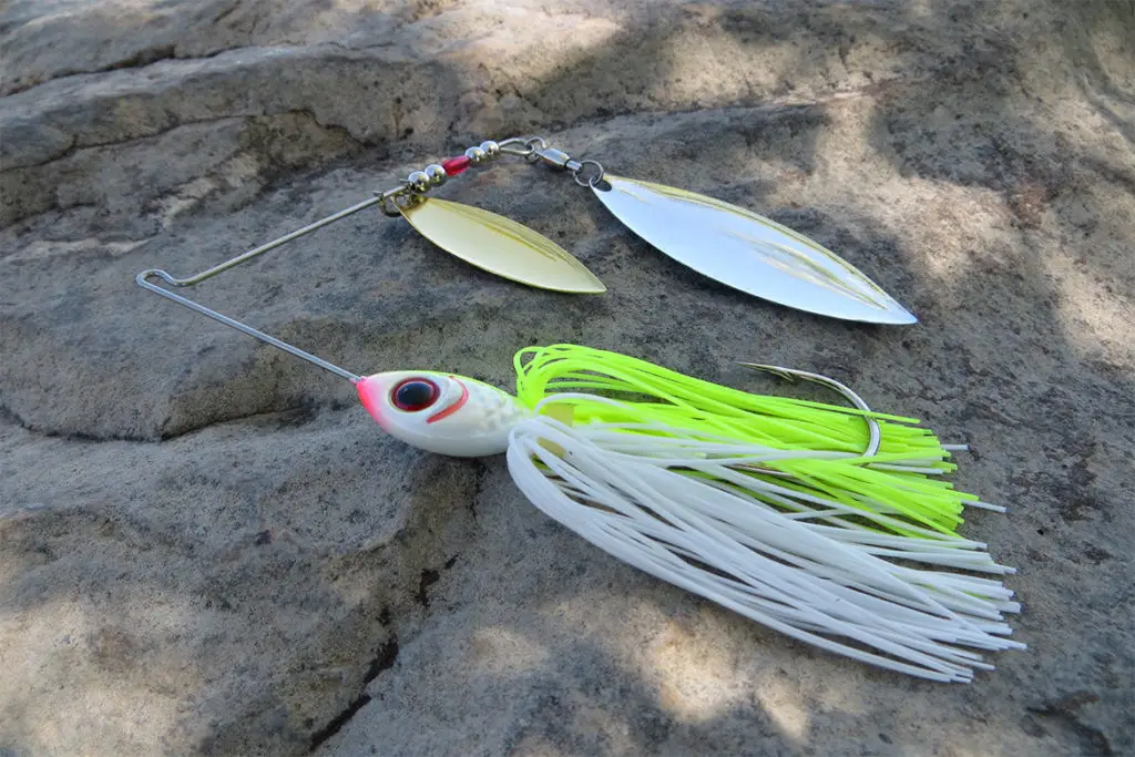 picture of a white and chartreuse spinnerbait sitting on a rock near a pond 